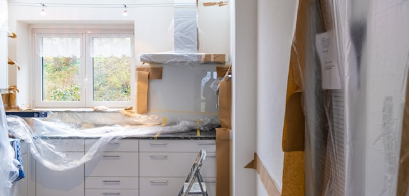 Kitchen under renovation with white cabinets, a window letting in natural light, plastic covers over surfaces, and a small ladder in the center.