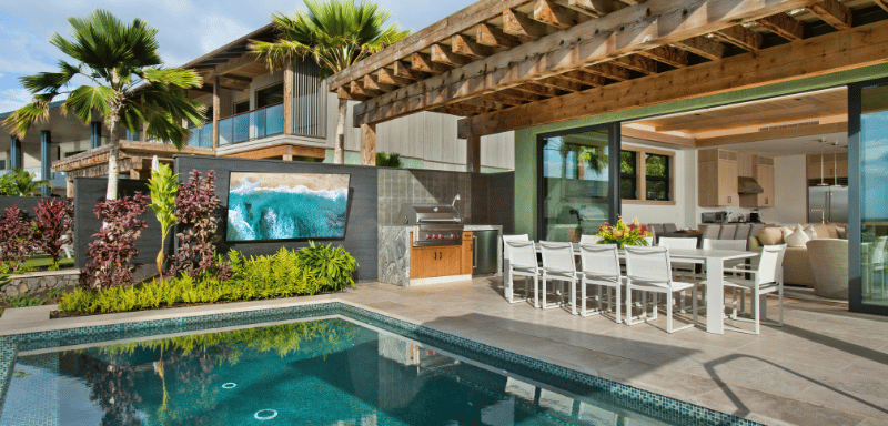 Modern outdoor living space with a swimming pool, covered patio with a large dining table and white chairs, outdoor grill, and open-concept kitchen connected to the living room.
