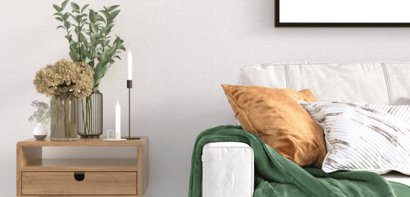 Close-up of a modern living room corner with a light beige sofa, orange and striped pillows, a green throw blanket, and a wooden side table decorated with vases, plants, and candles.