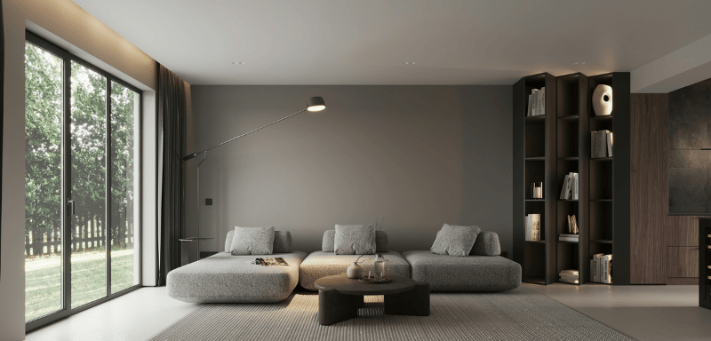 Modern minimalist living room with a large gray modular sofa, round coffee table, wall-mounted floor lamp, and a tall built-in bookshelf with books and decor items, next to wide glass doors overlooking a garden.