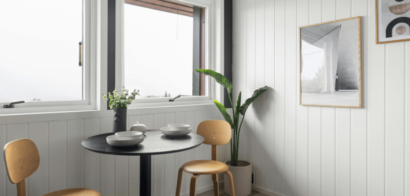 Small dining nook with a round black table set with two bowls, light wooden chairs, a tall green plant in a white pot, and wall art, next to large windows with natural light.