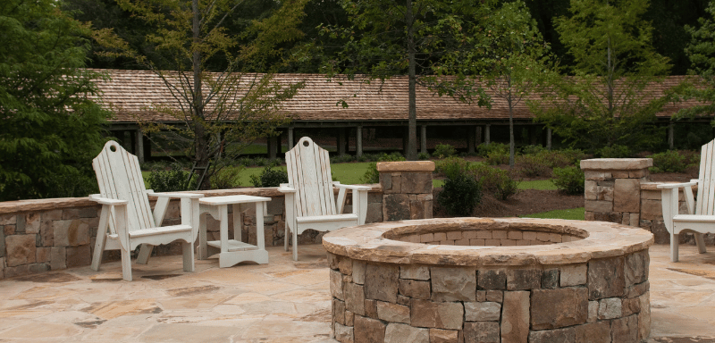 Stone patio with a circular fire pit surrounded by white Adirondack chairs and side tables, set in a landscaped garden with trees and greenery.