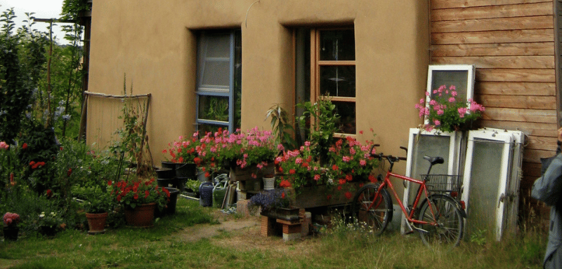 Rustic house with clay-colored walls and wooden siding, surrounded by potted flowers and plants, with a red bicycle parked near old window frames.
