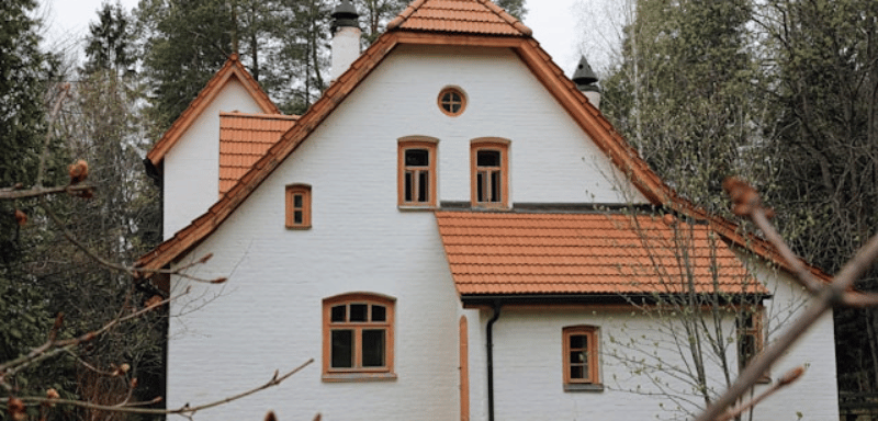 Two-story white brick house with multiple windows, a steep red tiled roof, and wooden trim, surrounded by tall trees.